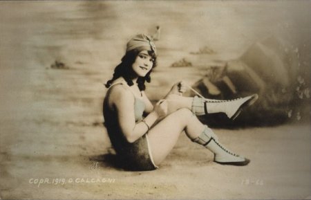 [hand-colored photographic image by D Calcagni from 1919 of young woman in bathing suit, sitting in a studio facsimile of a beach, lacing her foot-wear]